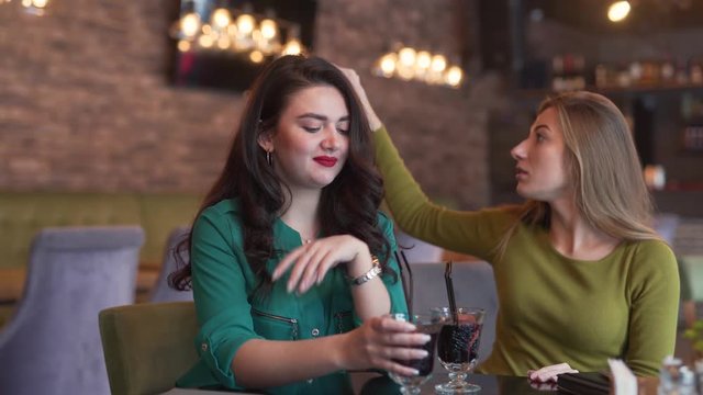 Two young women are talking in a video chat with their friend. One woman is drinking cocktail and other is making a video of a cafe by a phone. Both women is waving to third friend in the phone.