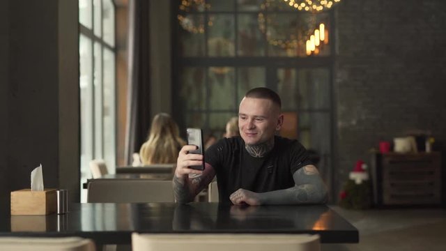 Young man with many tattoos on his neck and arms in a black t-shirt is sitting in a cafe indoors and talking on the video chat in his phone. Man is looking pleased and smiling a lot.