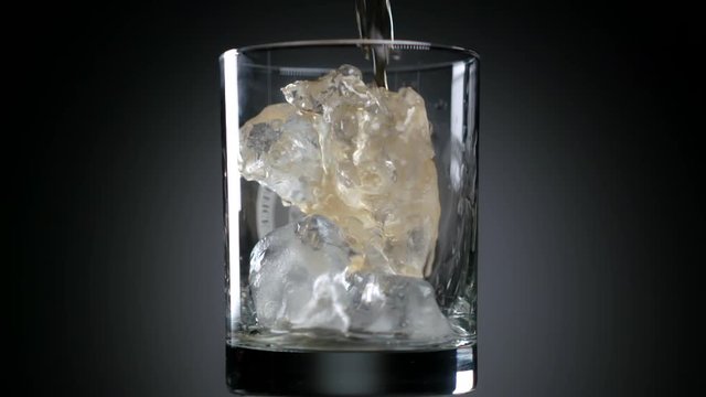 Whiskey or rum poured in super slow motion into a glass with ice cubes against gray background 