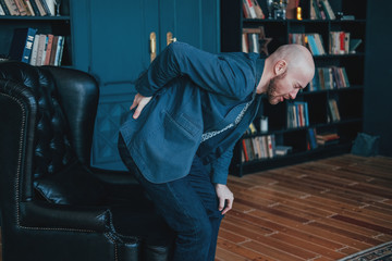 The attractive bald bearded man is holding onto his back, trying to get up from his leather chair....