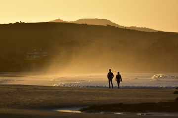 Fototapeta na wymiar Silhouette of couple walking on sandy beach next to white surf foam in yellow warm back light on sunset.