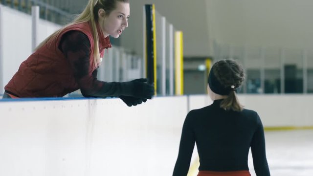 Professional figure skating trainer waiting for girl on skate near barrier inside ice arena to tell her comment about exercise