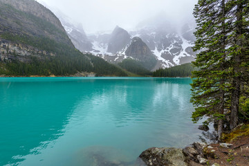 Fototapeta na wymiar Spring Moraine Lake - Spring view of snowy and foggy mountains reflecting in clear and emerald-color glacial water of Moraine Lake, Banff National Park, Alberta, Canada.