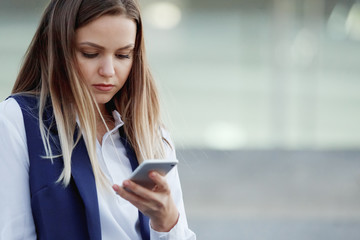 Young pretty woman with smartphone on the city street on the background of the sun at sunset. Looks at the phone.