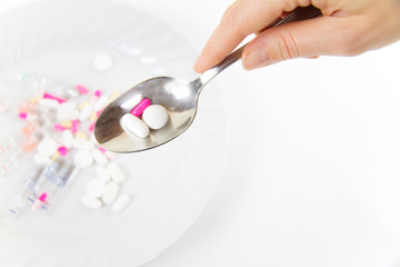 Addiction to medicines and drugs in modern times: a hand of a young woman with a spoonful of tablets taken from a plate full of medicines on white table and background
