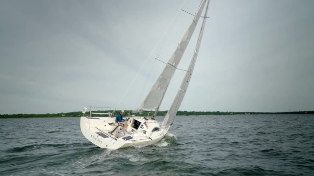 Man on his sail boat sailing on the ocean.  Climbs forward to adjust a sail. Shot from chase boat