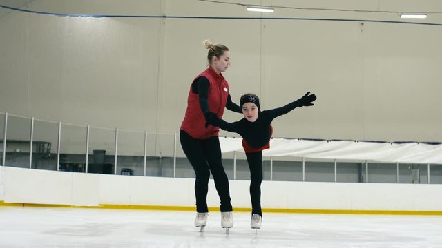 Young figure skating trainer moving near trainee, who making arabesque on ice inside arena, and helping to swing a leg right