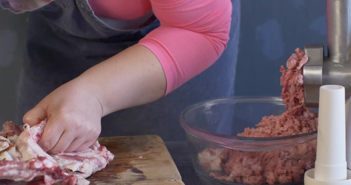 Slim woman cook in pink sweater, grey apron and white cap splits the leg of pig on wooden board. Grinds small pieces of meat with meat grinder and sorts minced meat into packages.