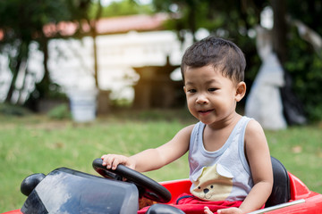 Little boy driving car with the steering wheel. Boy in a white shirt in a red toy car in the street. Little boy driving big toy car and having fun, outdoors.
