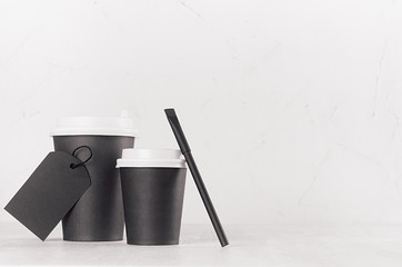 Coffee mockup - big, small black paper cups with white caps, blank label and sugar bag on white wood table with copy space, coffee shop interior. 