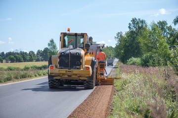 Road construction workers repairing highway road on sunny summer day. Loaders and trucks on newly made asphalt. Heavy machinery working on street. Road curbs being constructed with gravel 