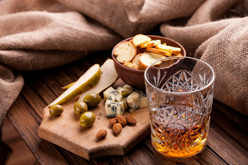 Rustic still life with whiskey and snacks. Crystal glass with a beautiful pattern on a wooden table. Snack for whiskey on crumpled paper olives, crackers, cheese, almonds, pear, apple.