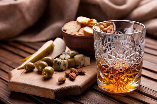 Rustic Still Life With Whiskey And Snacks. Crystal Glass With A Beautiful Pattern On A Wooden Table. Snack For Whiskey On Crumpled Paper Olives, Crackers, Cheese, Almonds, Pear, Apple.