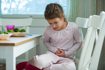 Child sitting at the table in the kitchen with stomach pain. Hands on belly. Little girl suffering