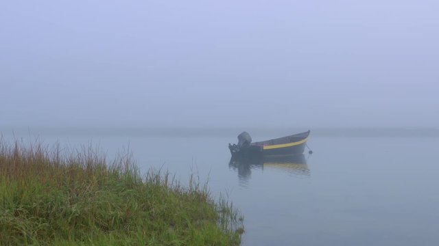 Black and yellow dinghy fishing boat at anchor in a quiet foggy cove with calm water and dune grass.