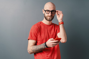 Handsome hairless Caucasian man with beard, glasses, red T-shirt talking on the phone on gray studio background