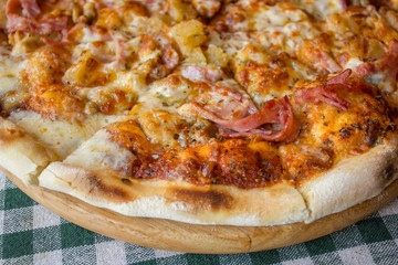 round pizza ready and cut pieces on a wooden plate on a checkered tablecloth close up