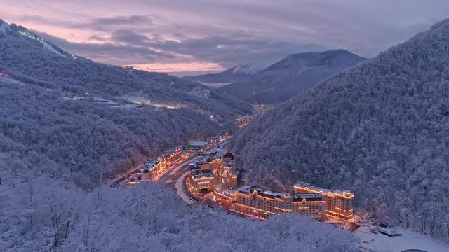 Aerial drone establishing shot of white winter valley Rosa Khutor resort in Sochi. Snow covered trees. Evening illumination. After sunset. Flying low above snow forest
