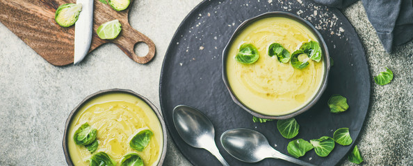 Flat-lay of seasonal brussels sprouts vegetable cream soup in black bowls and fresh green brussel sprouts on wooden board, top view, wide composition. Vegan, vegetarian, healthy, dieting food concept