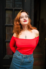 Fototapeta na wymiar Attractive redhaired woman in eyeglasses, wear on red blouse and jeans skirt posing at street against old wooden door.