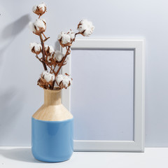 Mock up white frame and dry cotton twigs in blue vase on book shelf or desk. Minimalistic concept.