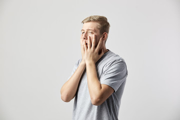 Sad guy dressed in a white t-shirt holds his hands on his face on the white background in the studio