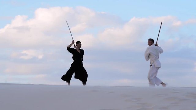 Concentrated men, in Japanese clothes, are practicing martial arts with a traditional Japanese weapon - a katana and jo,  in the desert at sunset.
