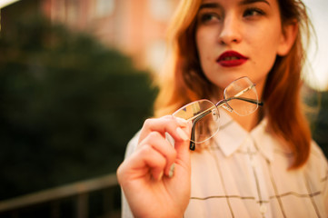 Close up photo of eyeglasses holding redhaired  woman, wear white blouse.