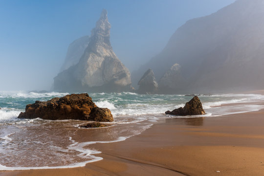 Fantastic seascape with sharp cliffs covered in fog rising from the turquoise sea waters and a beautiful untouched golden sand beach on a hazy spring morning. Ursa beach, Sintra, Portugal.