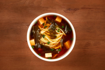 An overhead photo of a bowl of miso shiru soup with tofu, scallions, noodles, and wakame seaweed on a rustic wooden background with a place for text