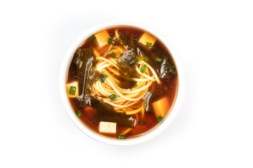A photo of a bowl of miso shiru soup with tofu, scallions, noodles, and wakame seaweed, shot from the top on a white background with copy space