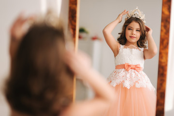 Little beautiful girl with brown hair in a Peach-colored dress. Girl try on diadem. Happy little princess look in to the mirror © Aleksandr