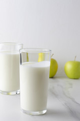 Vertical shoot of two glasses with fresh tasty kefir on white table in the kitchen, apples in the back