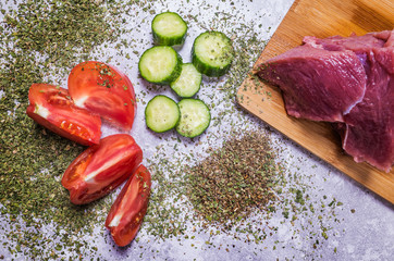 Products for cooking on a light background: a fresh piece of beef, sliced tomatoes and cucumbers, spices.