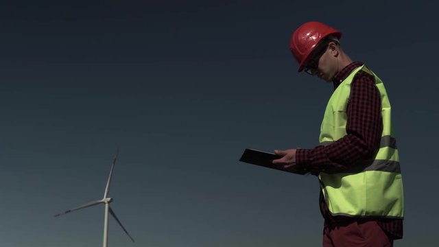 Engineer In The Green Vest With A Work Plan On The Background Of Windmills. Concept Of Alternative Energy