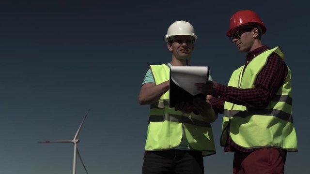 Two Young Engineers Men Approach A Windmill Power Generator And Discuss The Plan Of Its Repair In The Field On A Sunny, Clear Day