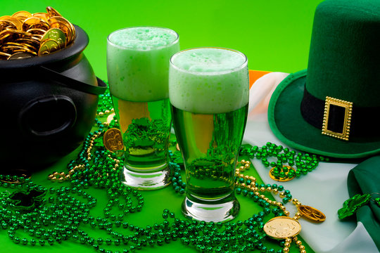 The luck of the Irish meme and Happy St Patricks day concept theme with two glasses of dyed beer, leprechaun hat, beads necklace and pot of gold coins on the Ireland flag isolated on green background