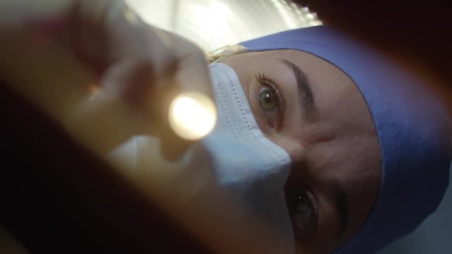 Close up shot of concentrated female doctor or nurse in surgical mask and cap using flashlight while examining patient before operation