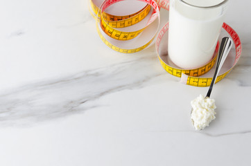 Yellow measuring tape,glass with milky product,spoon with cottage cheese on white marble table.Concept of milky diet and loss weight