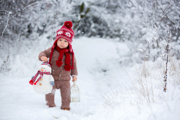 Fototapeta na wymiar Portrait of a cute toddler baby dressed in a brown hand knitted jacket, pants, red hat and scarf, holding teddy and lantern, walks through the snowy park enjoying first snow blowing