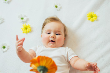 Beautiful baby lying on back reaching for a flower on white blanket 
