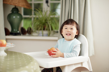 Fototapeta premium toddler girl eating apple sitting on high chair beside a dinner table at home
