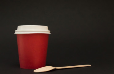red paper cup with a lid for coffee stands on a black background, next to it is a wooden coffee spoon