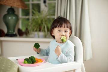 toddler girl eating  healthy  vegetable sitting on high chair beside a dinner table at home