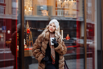 Outdoor close up portrait of beautiful  smiling woman blonde with curly hair, dressed in a white knitted hat and fur coat posing in street of european city. Winter fashion, Christmas holidays concept.