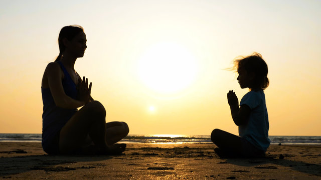 Silhouette of mother with little daughter meditating together at sunset