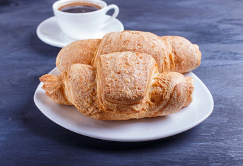 two croissants with cup of coffee on white plate on black wooden background