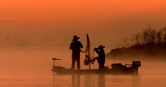 Two silhouetted traditional asian fishermen using nets to fish at an exotic river location from a flat-bottomed boat at sunrise or sunset