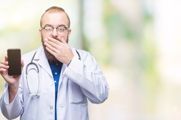 Young doctor man showing smartphone screen over isolated background cover mouth with hand shocked with shame for mistake, expression of fear, scared in silence, secret concept