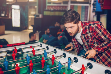 Young man playing alone at table soccer in room. Intense game. Guy looks concentrated.